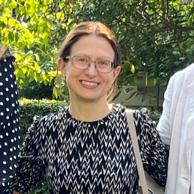 A woman with glasses and brown hair tied back, wearing a patterned black and white top, stands outdoors smiling in front of greenery.