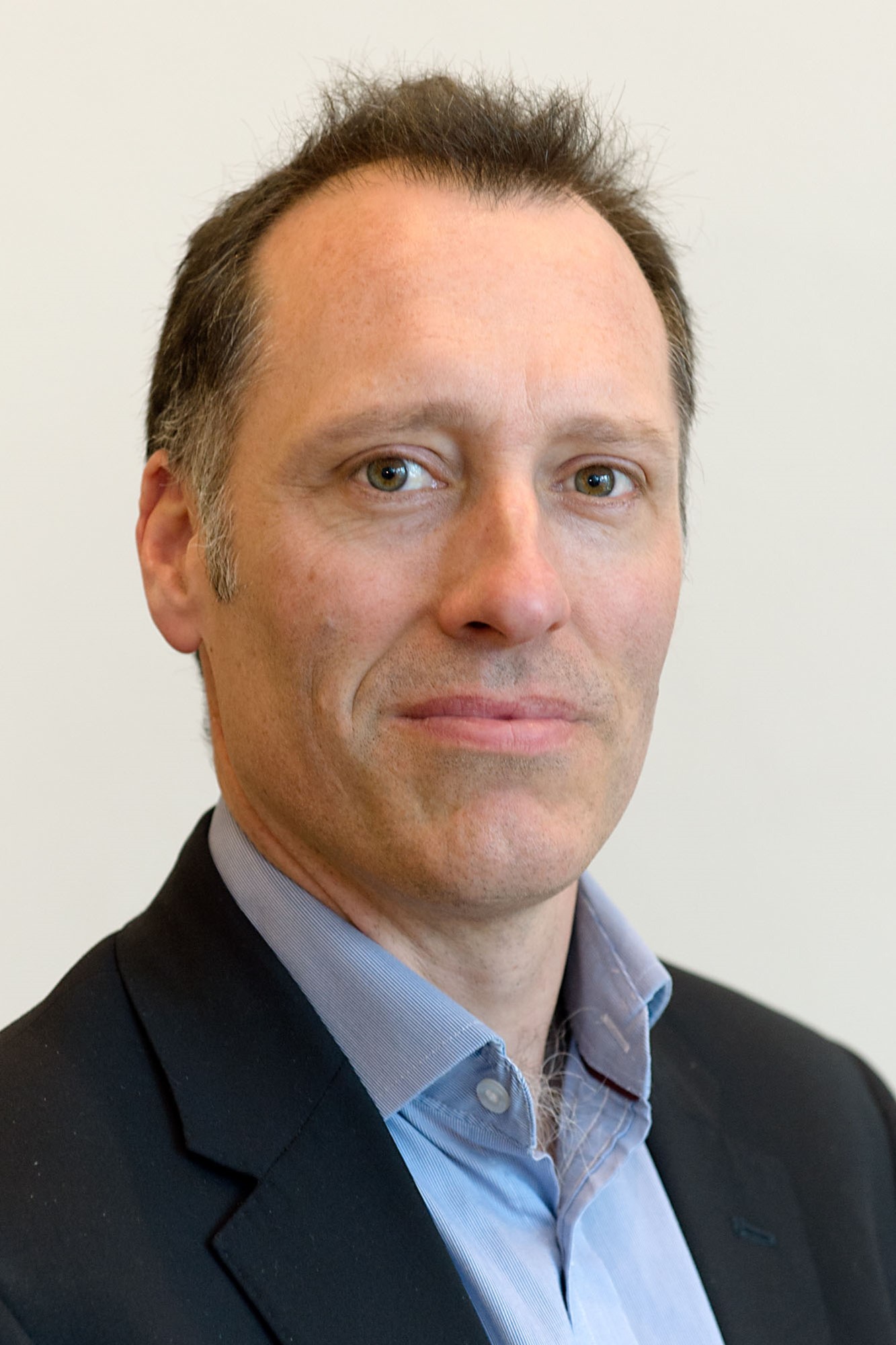 Professor Richard Adam - A middle-aged man with short brown hair wearing a dark jacket and light blue shirt stands against a plain light background, facing the camera with a neutral expression.