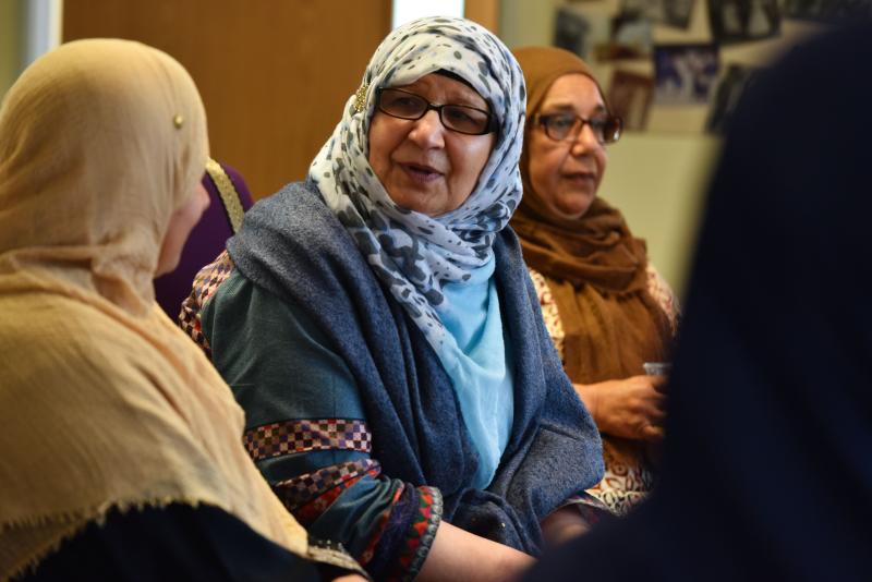 Three women wearing headscarves sit together indoors, engaged in conversation. The focus is on the woman in the center, who is smiling and wearing glasses.