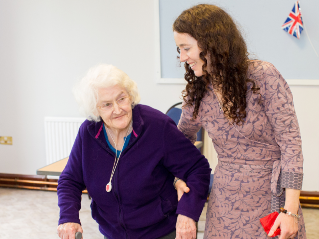 Elderly woman walking with frame linking arms with younger woman