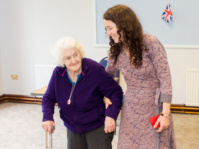 Woman wearing a dress walking an older woman with a walking stick