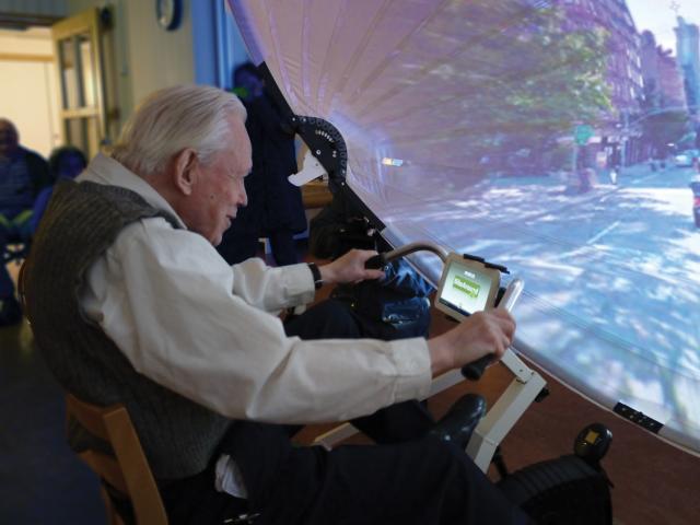 Grey haired man cycling on exercise bike in front of screen showing a street view