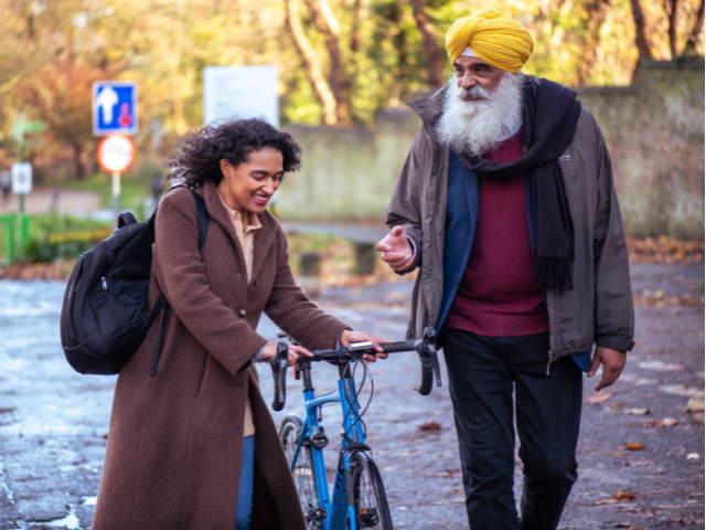 Woman walking with bike and talking to man with grey beard and yellow turban