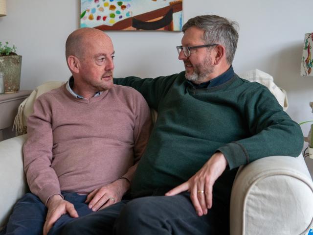 Two men sitting on an armchair together, one is wearing and pink jumper and is bald, the other is wearing a green jumper and has grey hair and glasses. They are looking at each other with a serious expression. 