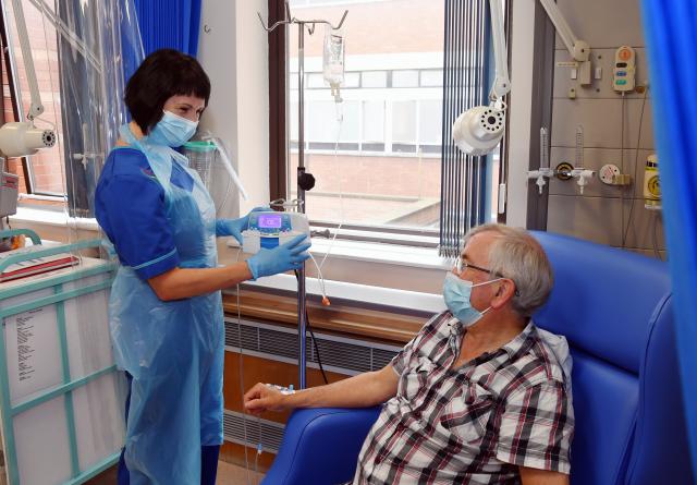 A female nurse with a mask and dark hair on left hand side talking to a male patient receiving chemotherapy.
