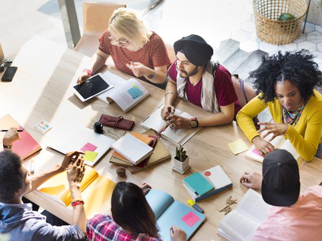 A group of six young students sitting around a table with open notebooks.