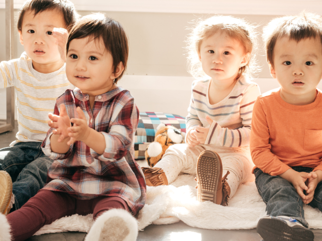Four toddlers sitting on a floor and clapping.