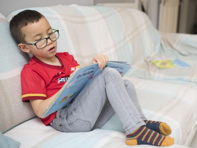 A boy reading a book alone on a sofa. 
