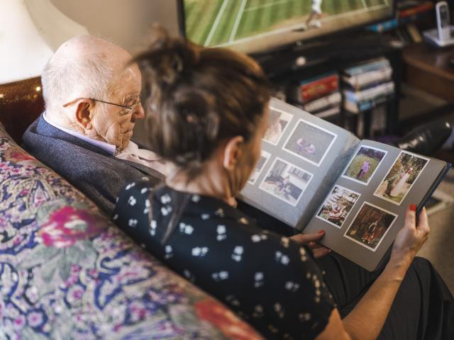 A woman and an older man sitting on the couch and looking at a phot album. 
