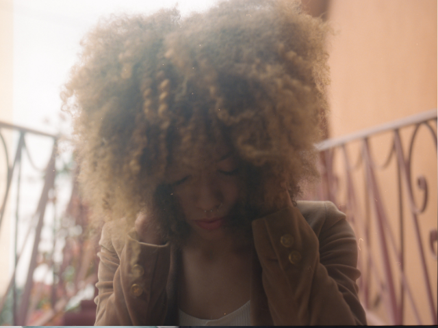 A young black woman with covering her face with her hair and holding her head in her hands. Her eyes are closed and looks in deep though.