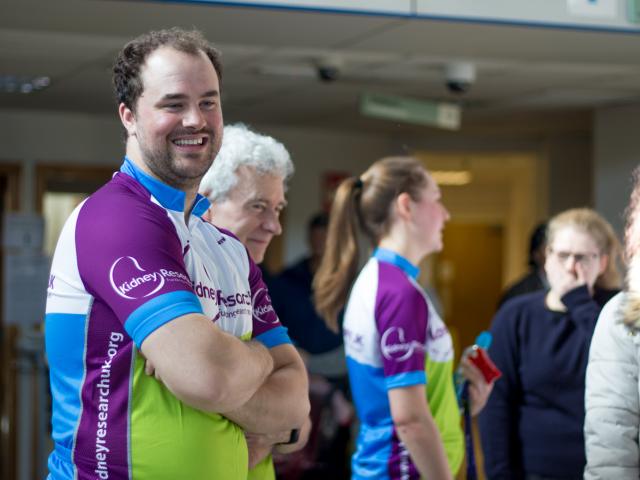 A man representing wearing a purple, green and white t-shirt with Wales Kidney Research logo is smiling at the camera.