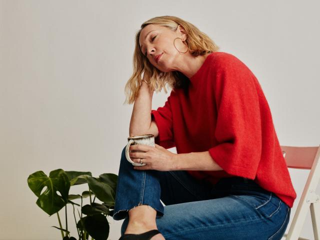 A woman with short blonde hair and bright red sweater on is sitting on a chair. She is holding a cup and there is a green plant on the left to her.