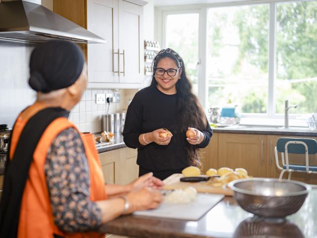 A younger dark-haired woman is in the kitchen smiling down at potatoes. Opposite her an older woman is chopping onions.