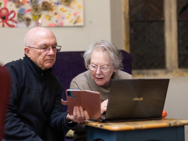 An older couple, a woman and a man, struggling to use technology aka a smartphone and a laptop.