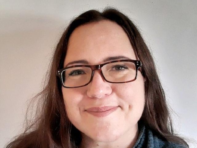 A headshot of Zoë Abbott: a woman with long brown hair and glasses with a smile on her face..