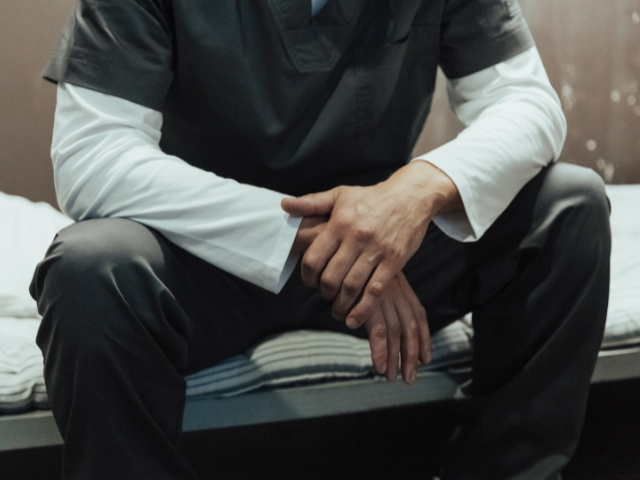 Person in dark scrubs with a long-sleeved white shirt, sitting on a bed, hands clasped and resting on knees, in a dimly lit room.