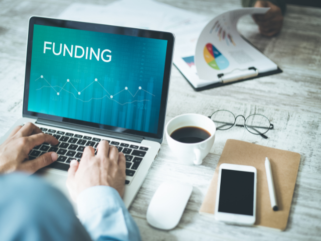 Person typing on a laptop displaying the word "FUNDING" and a graph. Nearby are a smartphone, coffee cup, mouse, and a folder with papers. Another person is holding a document.