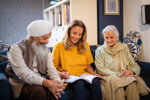 Three people sit on a couch, one in a white turban, one in yellow writing in a notebook, and one elderly woman in a light shawl; books and decor are in the background