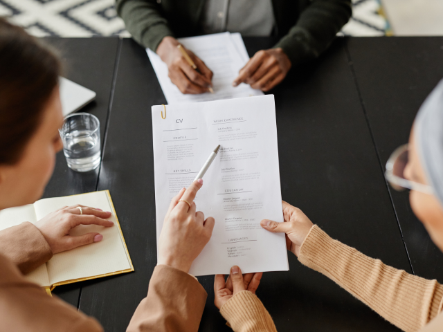 Three people sit at a table reviewing a printed CV. One person points at the document with a pen. A glass of water and a notebook are nearby.