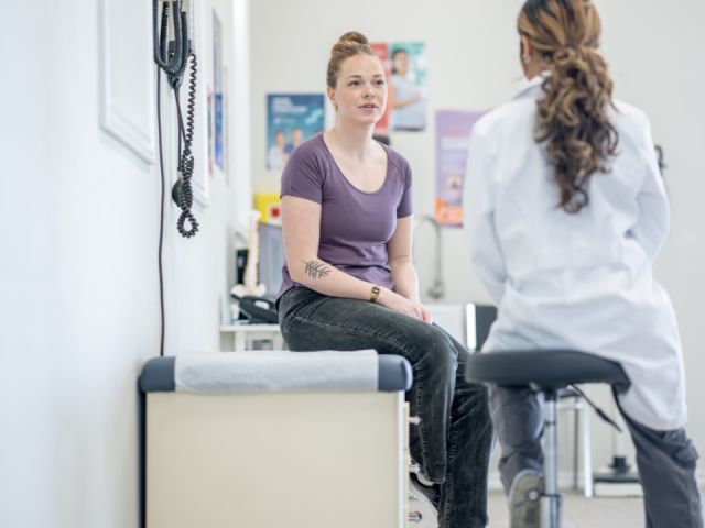 A woman in a purple shirt sits on an examination table talking to a healthcare professional in a white coat in a medical office setting.