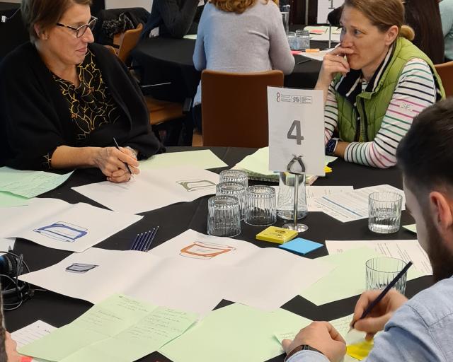 Three people sit around a table with papers, pens and glasses of water, engaged in discussion at a workshop. A sign with the number 4 stands on the table.