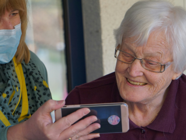 a_woman_wearing_face_mask_shows_elderly_woman_her_phone