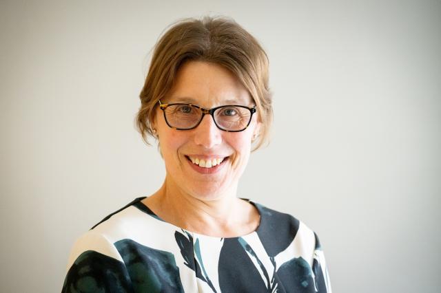 A woman with short brown hair and glasses is smiling, wearing a patterned top, and standing in front of a plain light-coloured background.