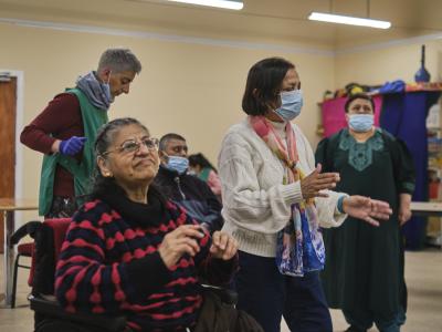A group of disable individuals dancing. The focal point of the picture are two women, one in a red sweater and in the wheelchair, and then other one standing right to her and clapping her hands. 
