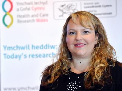 Dr Denitza Williams posing in front of Health and Care Research Wales branded wall.