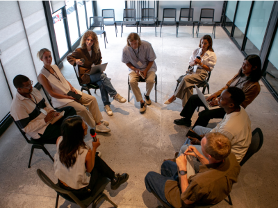 A group of people seated in a circle in a modern room, some holding laptops, engaged in discussion.