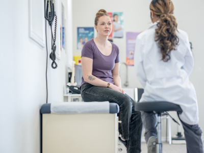 A woman in a purple shirt sits on an examination table talking to a healthcare professional in a white coat in a medical office setting.