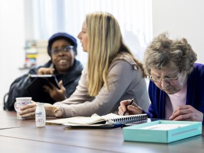 Three people sitting at a table in a meeting room. One woman focuses on documents, another woman speaks, and a person is smiling while holding a tablet.