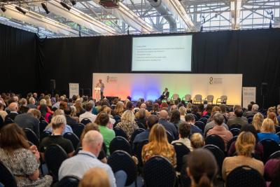 A large audience listens to a speaker presenting slides at a conference in a spacious, well-lit hall with rows of chairs and a stage.