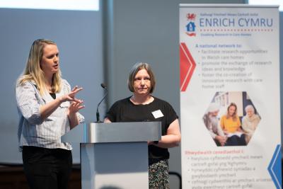 Two women stand at a podium speaking; a presentation banner about Welsh care home research is visible in the background.